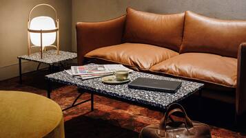 A cozy room with a leather sofa, a coffee table with a laptop and a cup, a lamp, and a handbag.