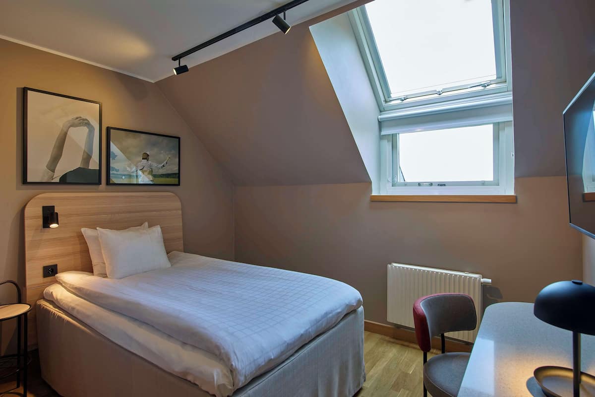 A bedroom with a bed, a desk, a chair, a radiator, and two framed pictures on the wall.