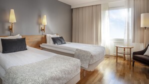 Quarto de hotel com duas camas, uma mesa pequena, uma cadeira e uma janela com cortinas.