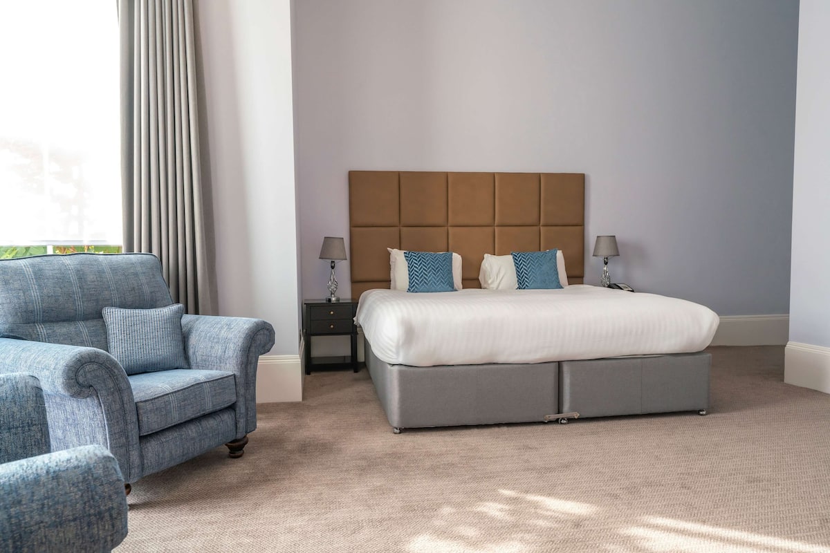 A bedroom with a bed, a chair, two nightstands, and a window with curtains.