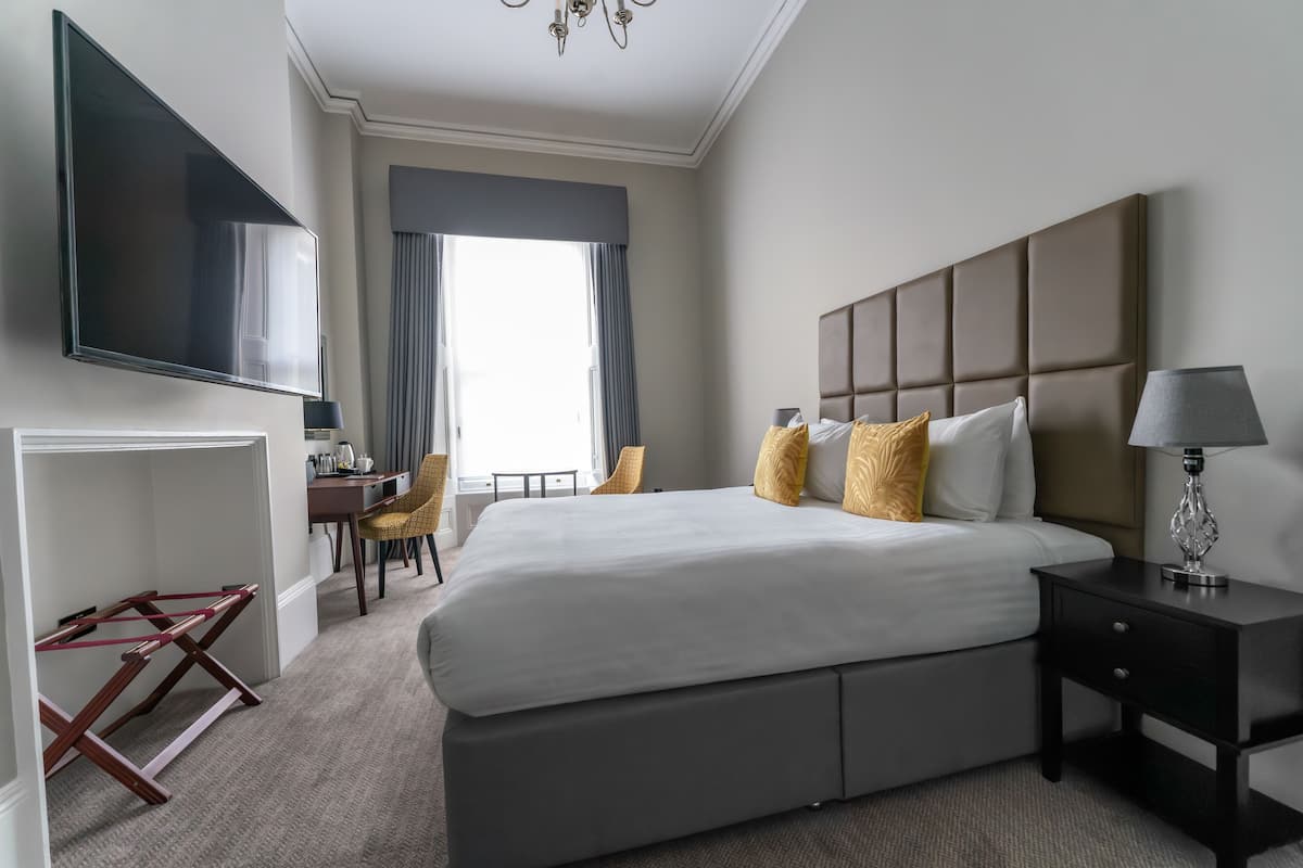 A modern hotel room with a large bed, a flat-screen TV, a desk with a chair, and a window with curtains.