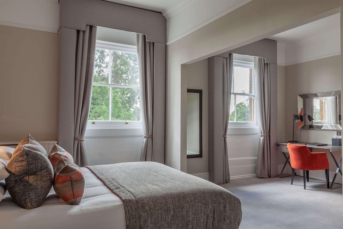 A bedroom with a bed, a desk, and a chair. There are curtains on the windows.