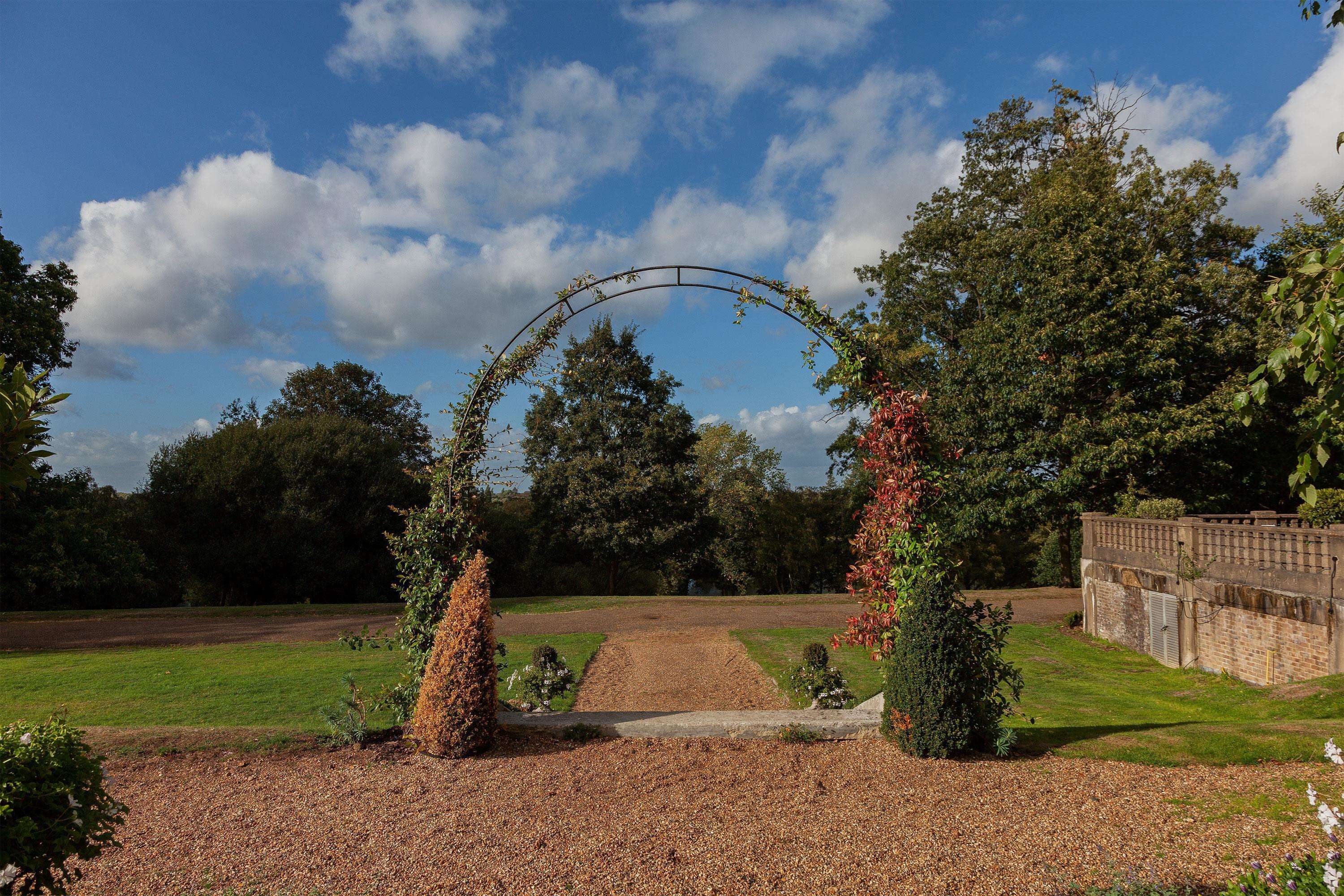 An arched pathway leading to a building with a brick wall and a garden with trees and shrubs.