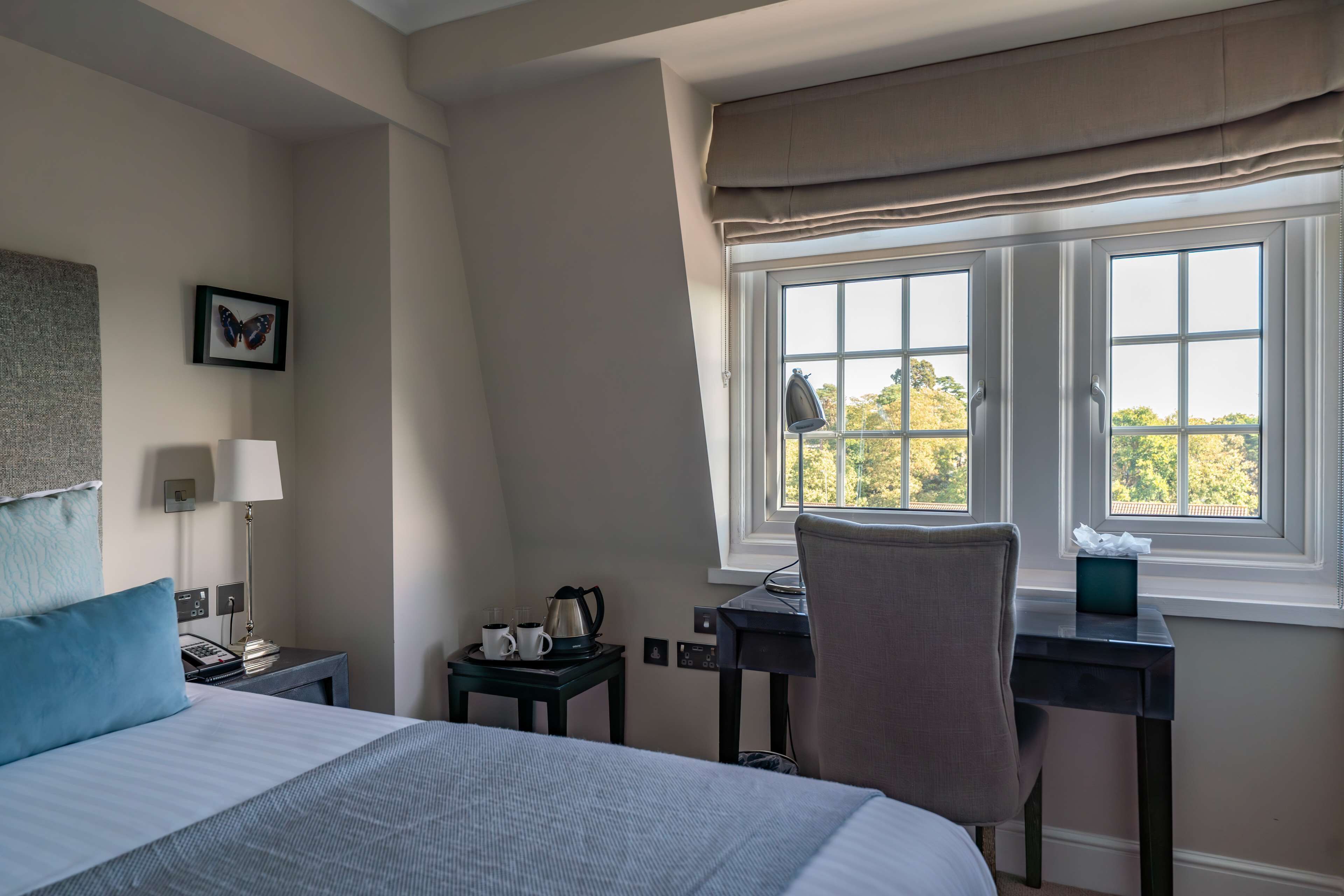 Une chambre d’hôtel avec un grand lit, un bureau avec une chaise, une fenêtre avec des rideaux, une table de chevet avec une lampe, un téléphone, une cafetière et un tableau représentant un papillon, accroché au mur.