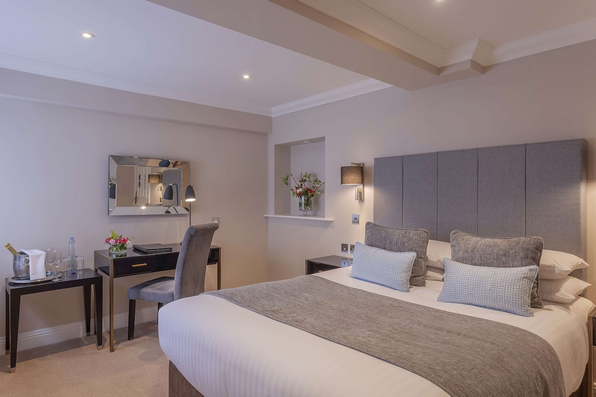 A modern hotel room with a large bed, a desk, a chair, a mirror, and a nightstand with a lamp.