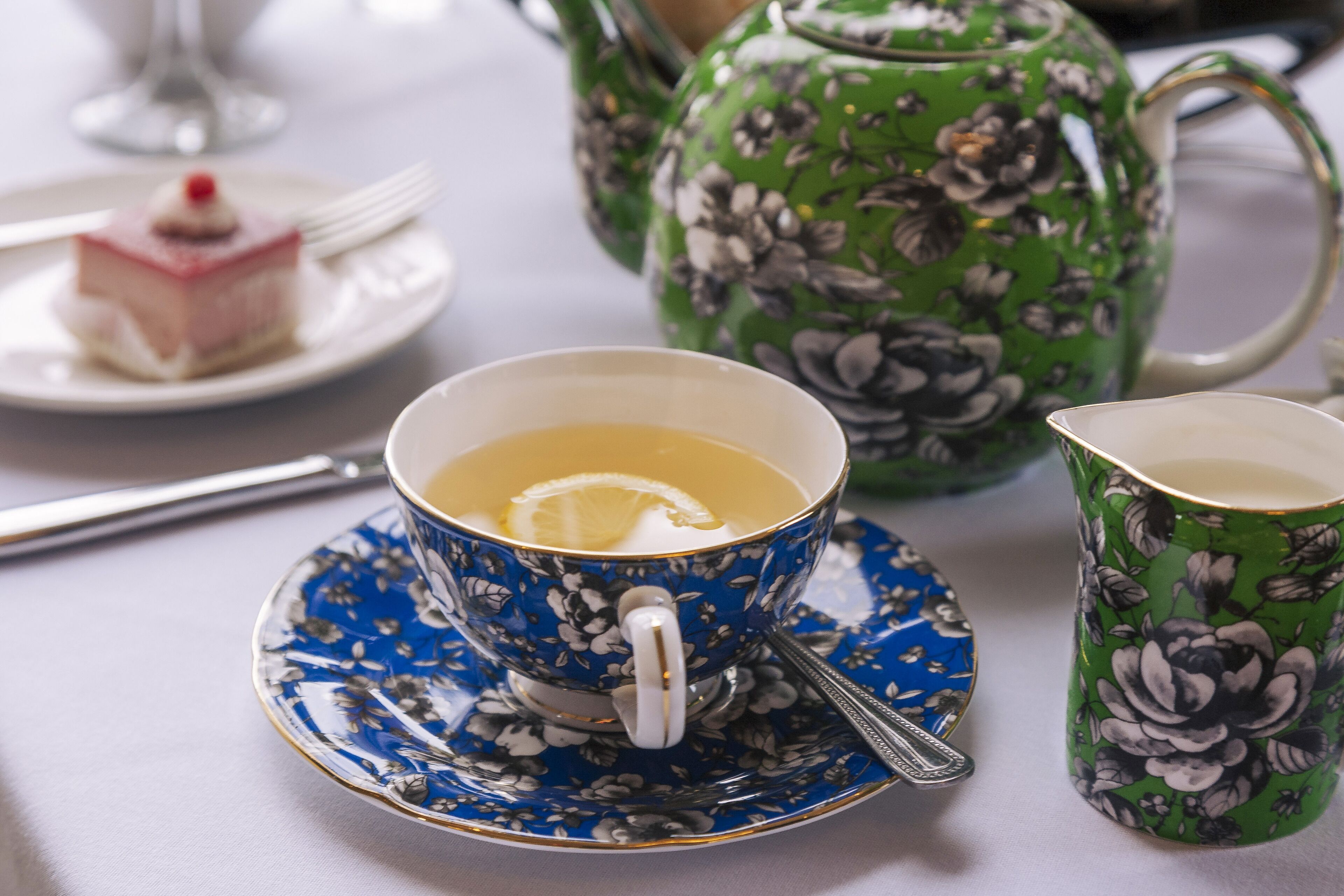 Une table dressée avec une théière à motifs floraux, une tasse de thé avec une tranche de citron et une petite pâtisserie sur une assiette.