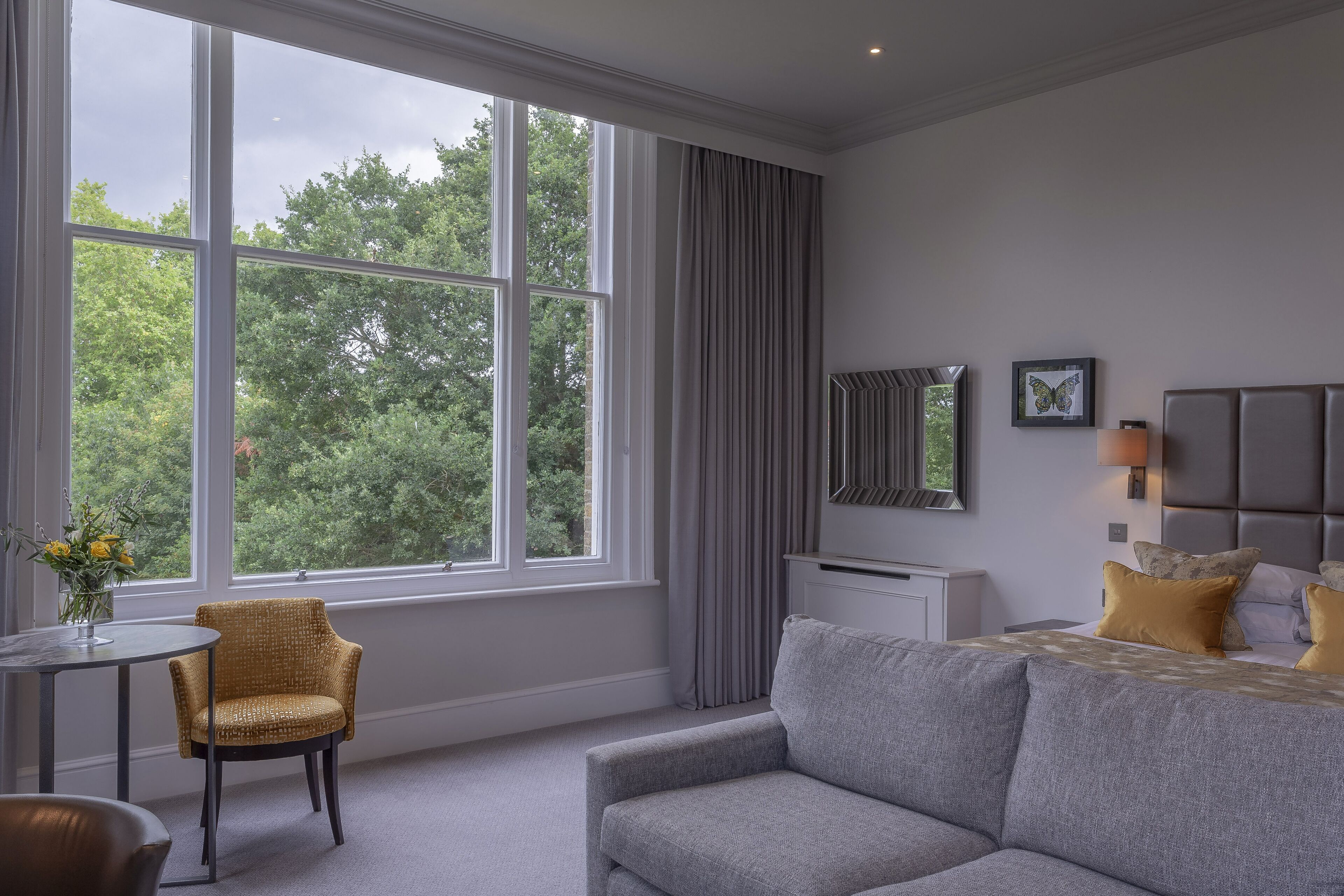 Une chambre d’hôtel moderne avec une grande fenêtre, un canapé gris, une petite table sur laquelle est posé un vase de fleurs, et un lit avec une literie grise et des oreillers jaunes.