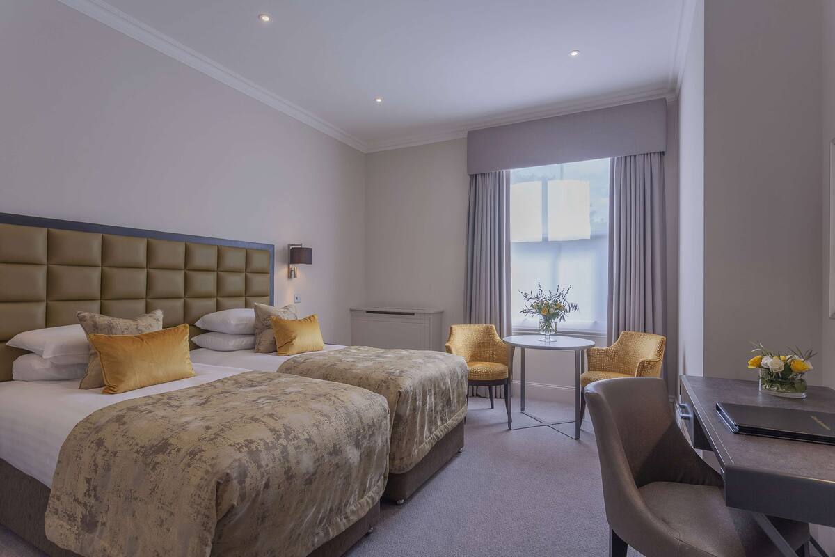 A hotel room with two beds, a small table, a chair, and a view of the city.