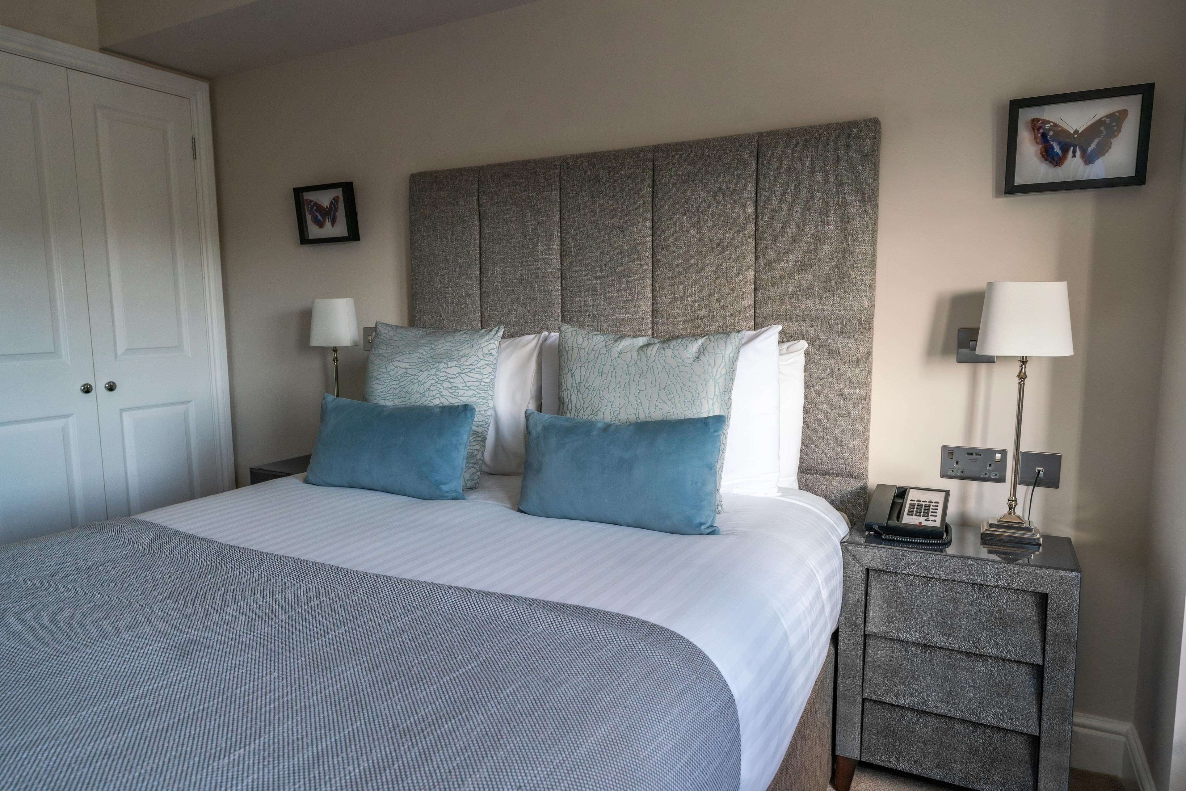 A neatly made bed with a grey and white striped bedspread, two blue pillows, a grey headboard, a bedside table with a lamp, and a framed butterfly picture on the wall.