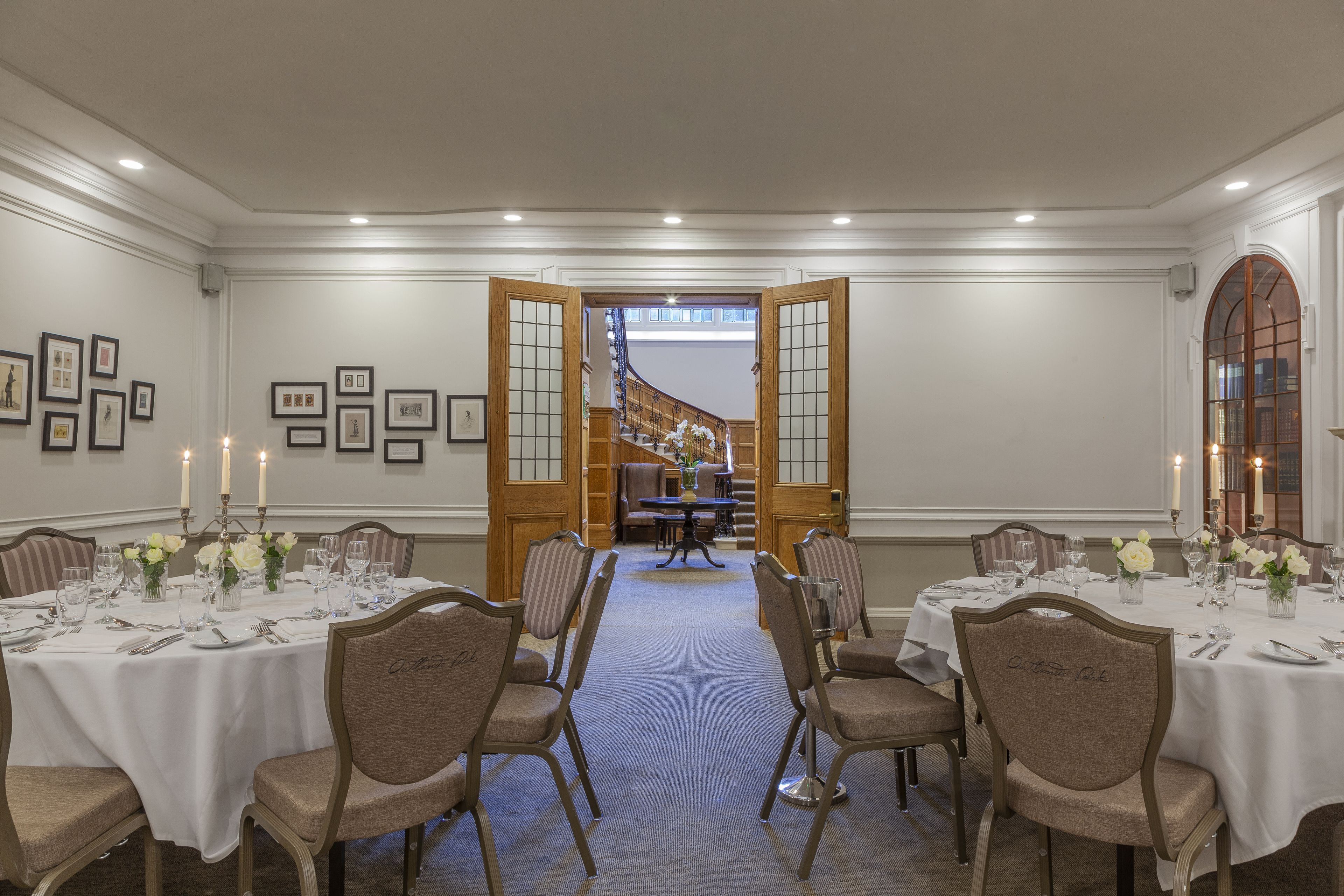 A formal dining room with tables set for six, each with white tablecloths, floral centerpieces, and candle holders. The room has a classic architectural design with wooden doors and a staircase in the background.