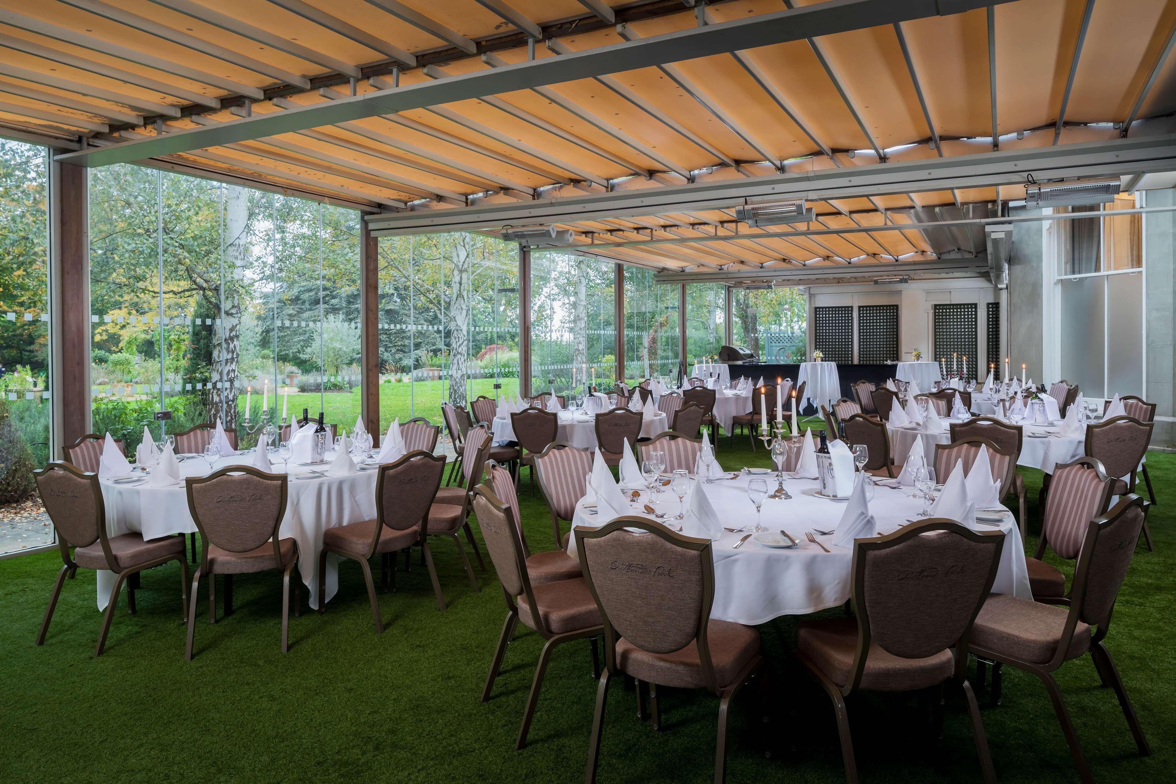 An outdoor dining area with round tables and chairs, covered by a wooden pergola, set against a backdrop of trees and a grassy lawn.