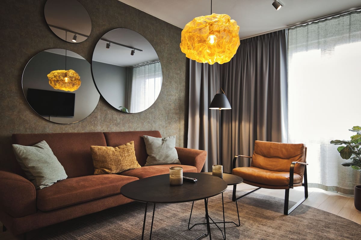 A modern living room with a brown sofa, a round coffee table, and a leather armchair. There are three round mirrors on the wall, a large yellow pendant light, and a black floor lamp.