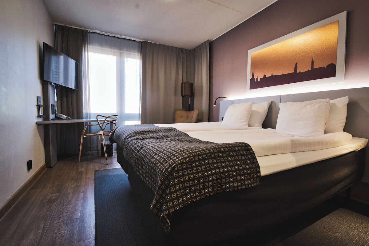 A hotel room with a bed, a desk with a chair, a television, and a painting on the wall.