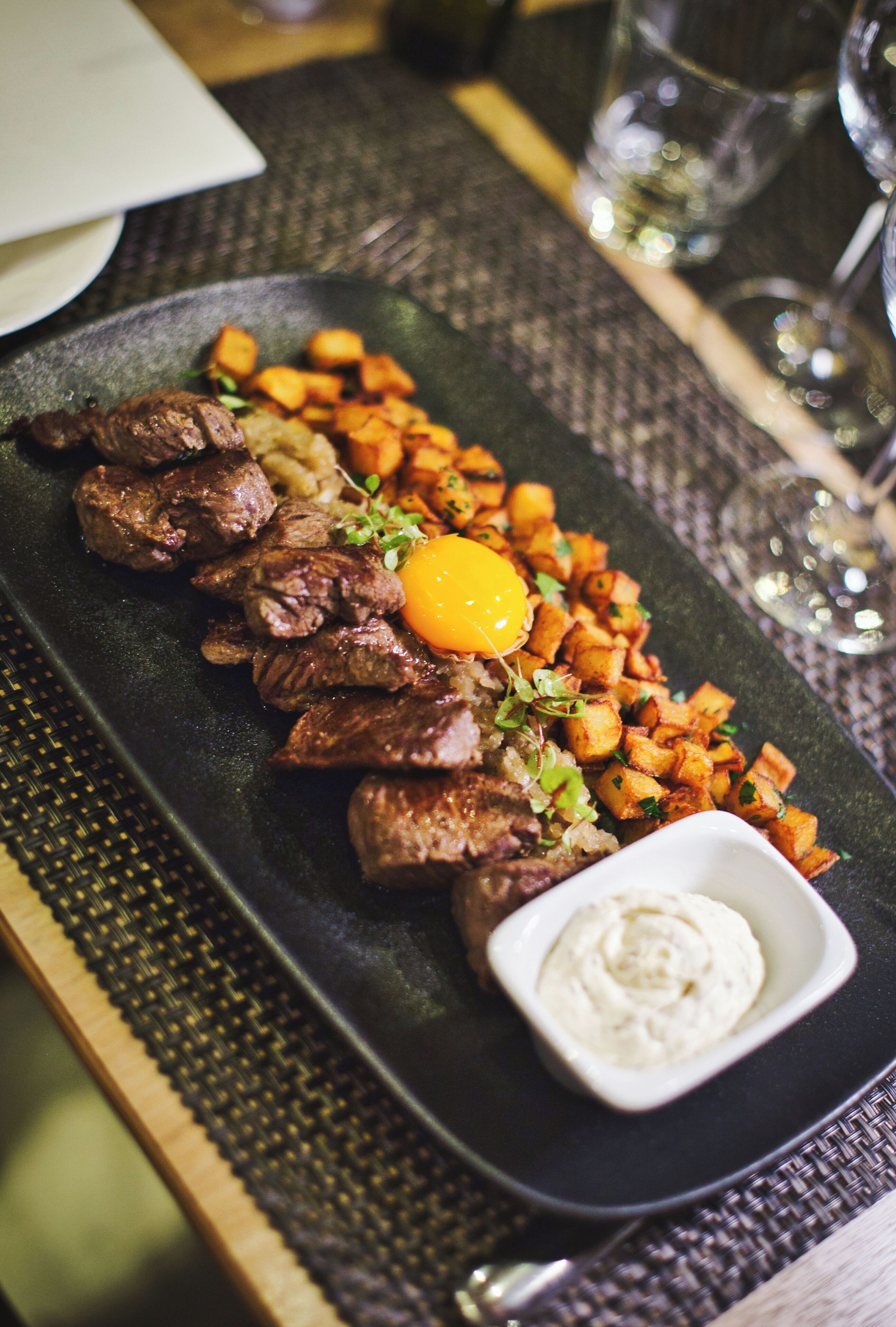 A rectangular plate with steak, roasted potatoes, and a raw egg yolk, accompanied by a small bowl of sauce, set on a table with a woven placemat.