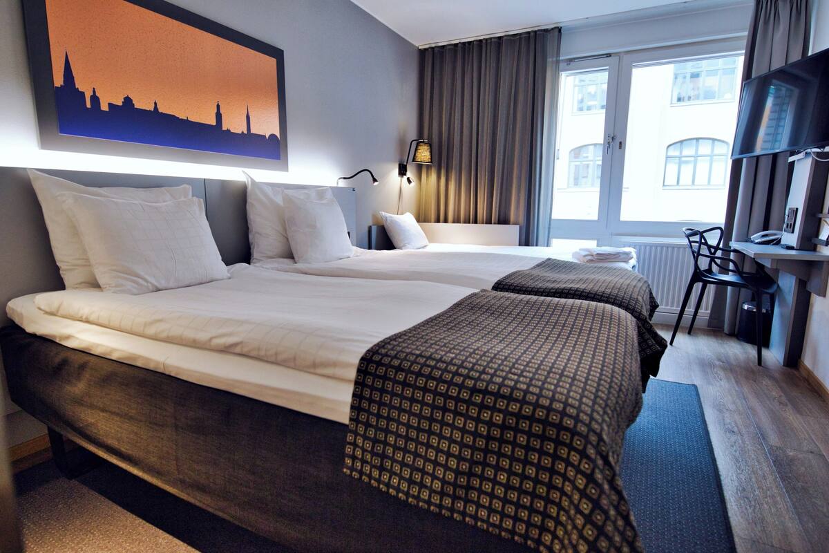 A hotel room with two beds, a desk, a chair, a TV, and a cityscape painting on the wall.