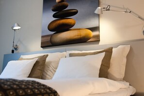 A neatly made bed with white and brown pillows, a wall-mounted lamp, and a poster of stacked stones.
