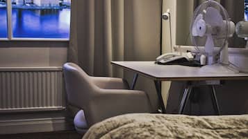A hotel room with a bed, a desk with a fan, a chair, and a view of a cityscape through a window.