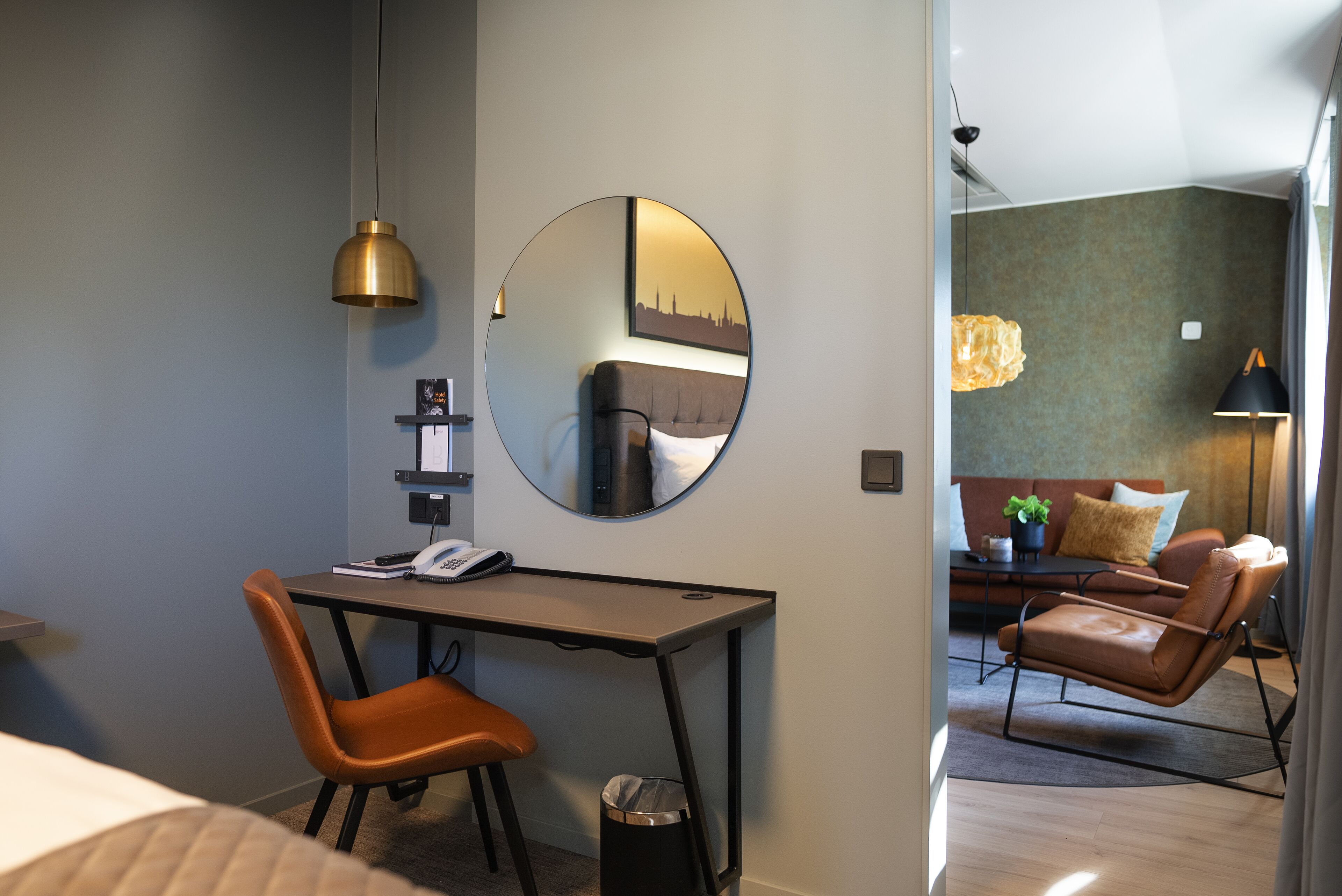 A hotel room with a desk, chair, and a large mirror. There is a sofa and a lamp in the adjacent living area.
