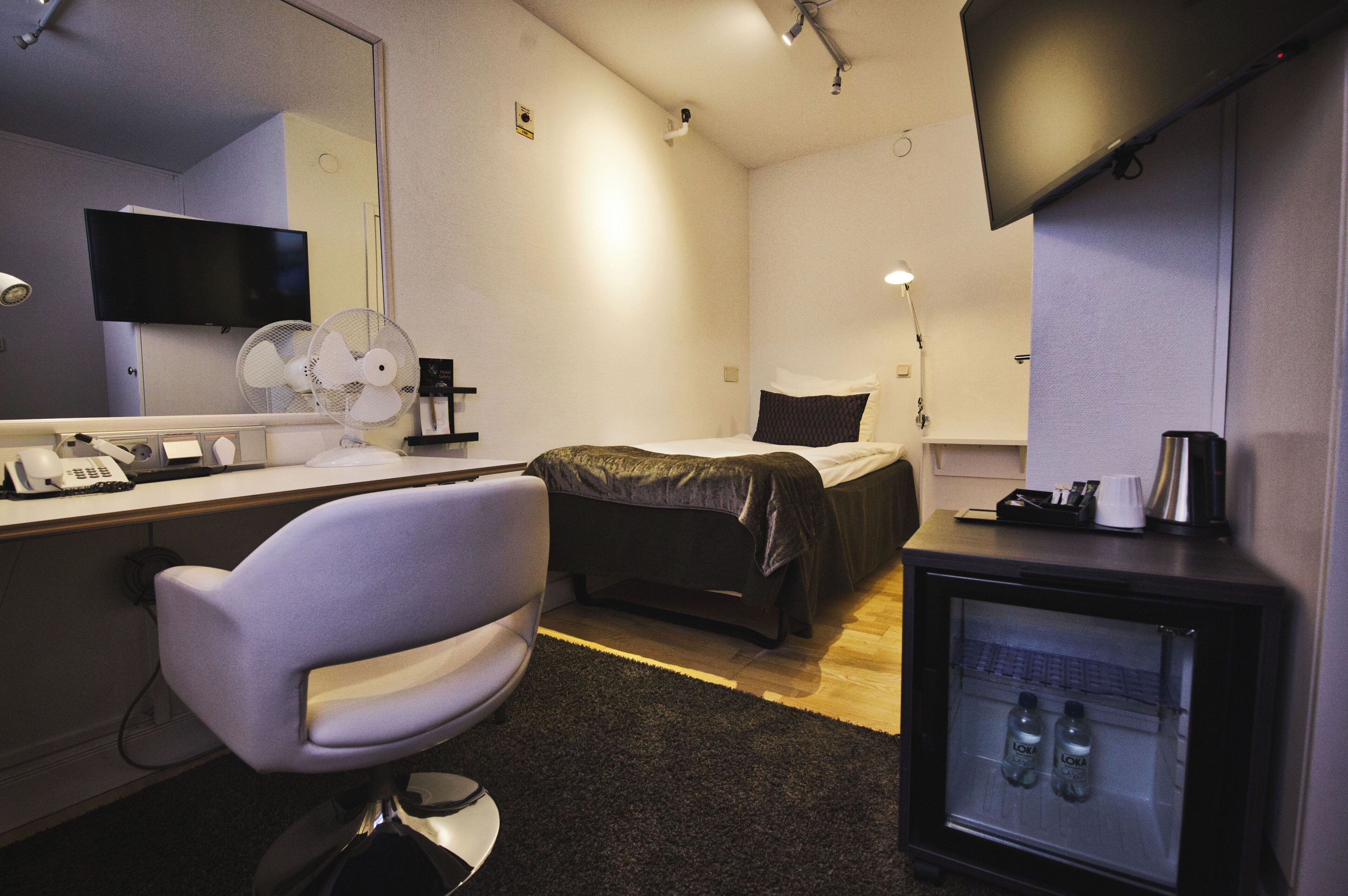 A hotel room with a bed, a desk with a computer and phone, a chair, a TV, a mini-fridge, and a mirror.