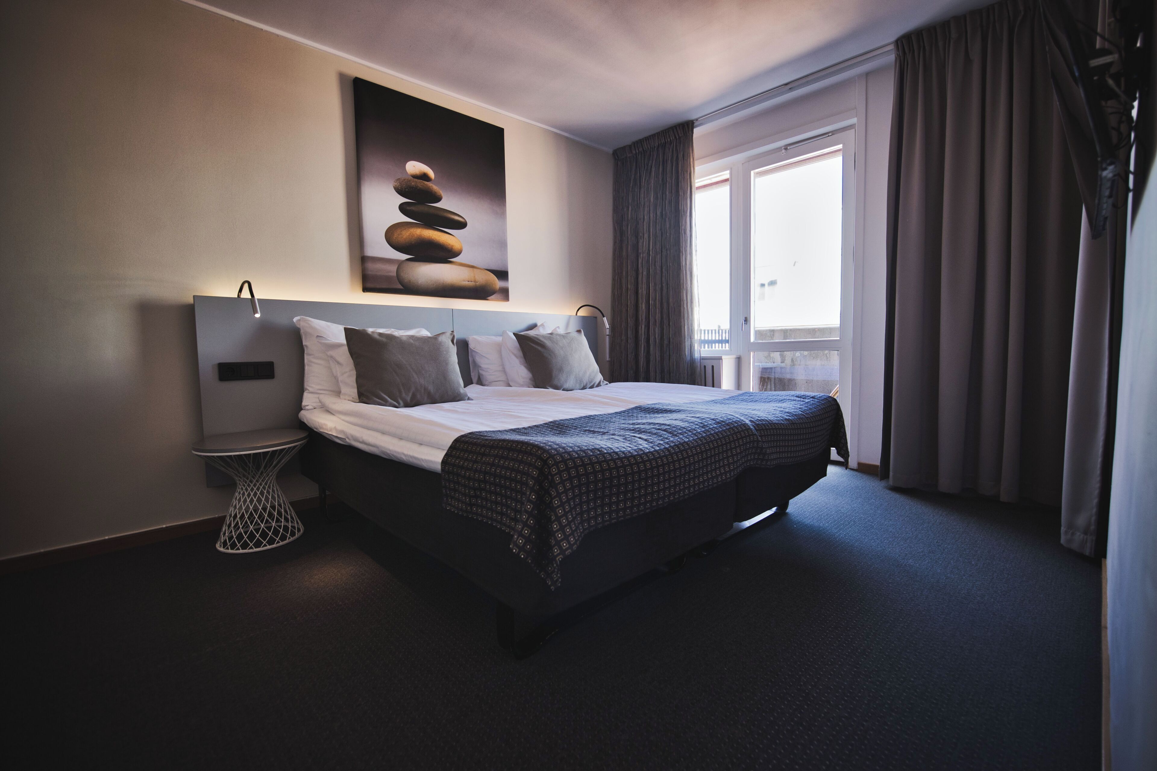 A modern hotel room with a large bed, a bedside table, a wall-mounted light, and a window with curtains.