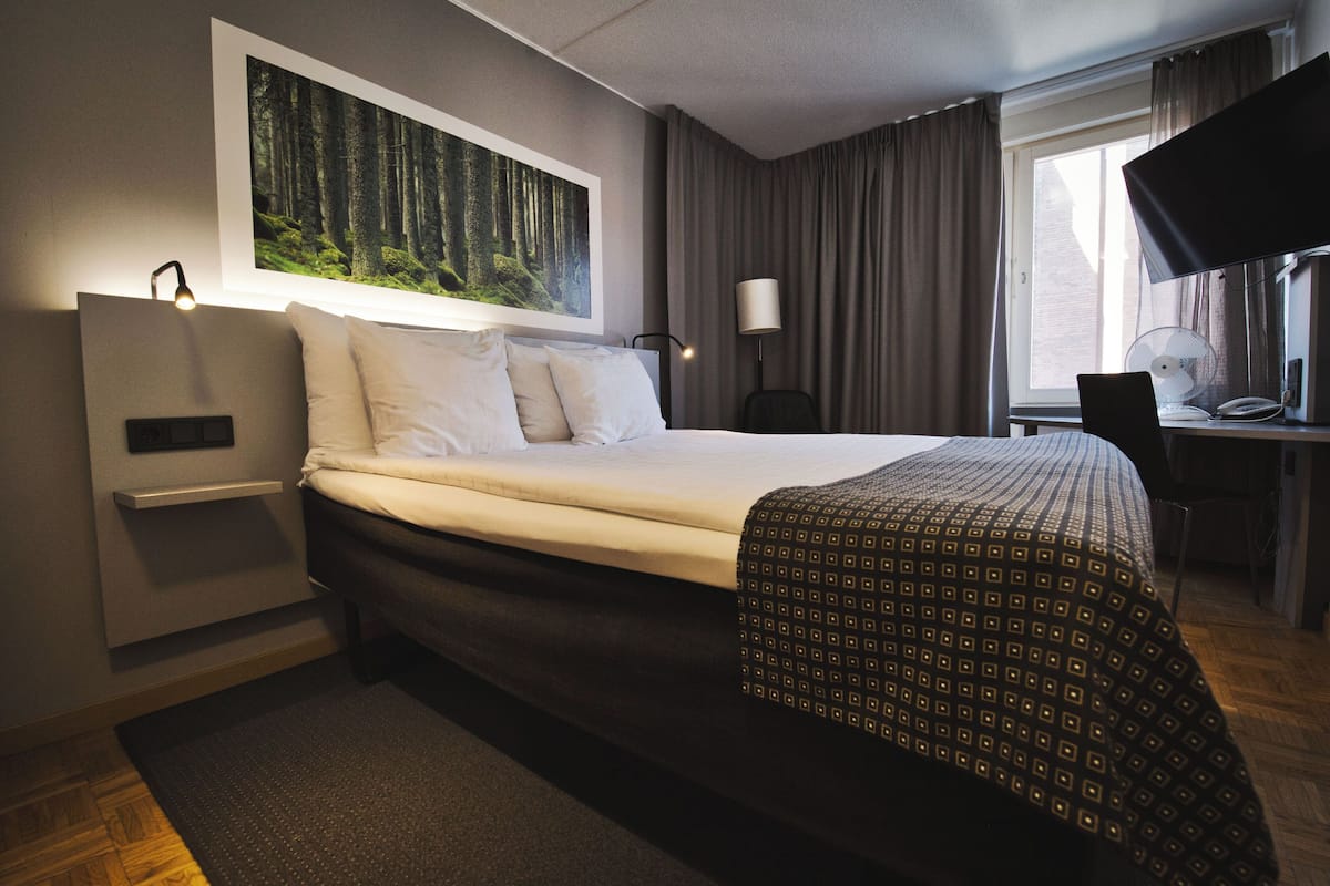 A hotel room with a bed, a desk with a computer, a chair, a desk lamp, a wall-mounted TV, and a large window with curtains.