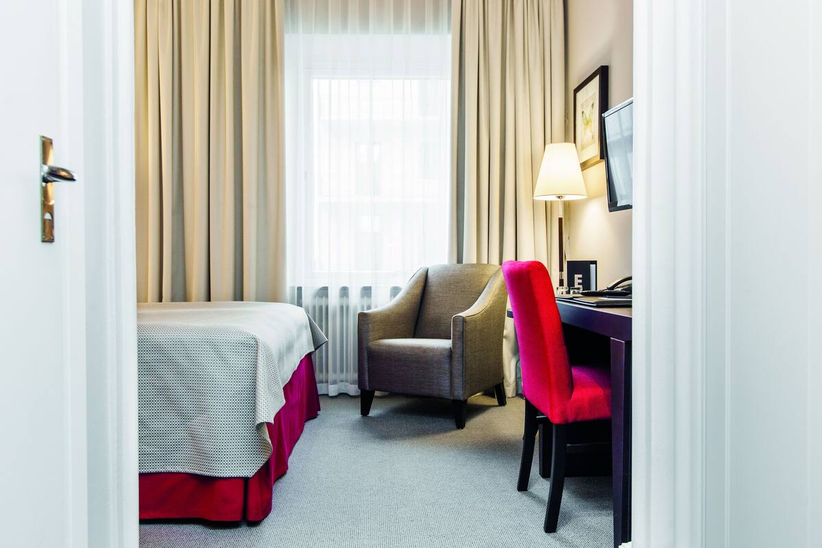 Una habitación de hotel con cama, escritorio, silla y ventana con cortinas.