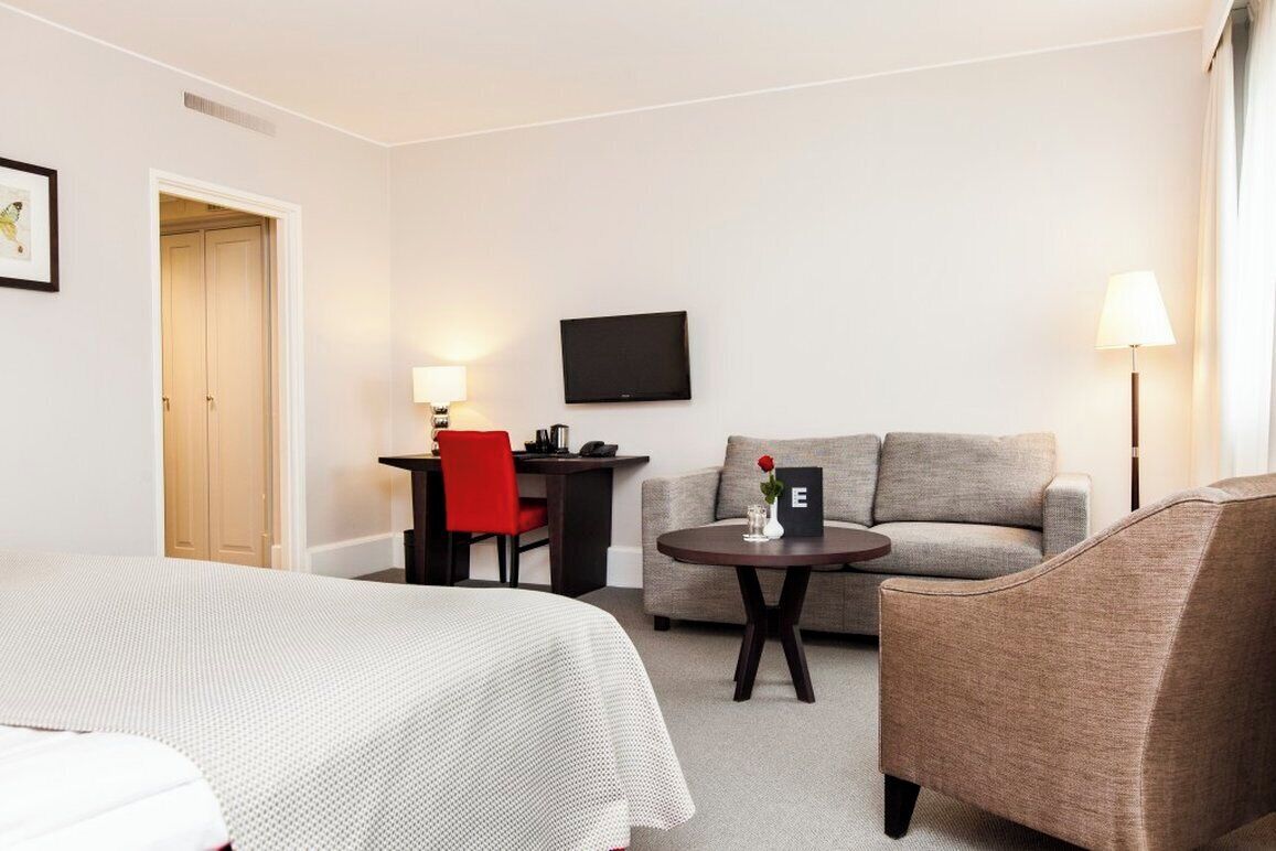 A hotel room with a bed, a sofa, a desk with a chair, a small table with a lamp, and a TV on the wall.