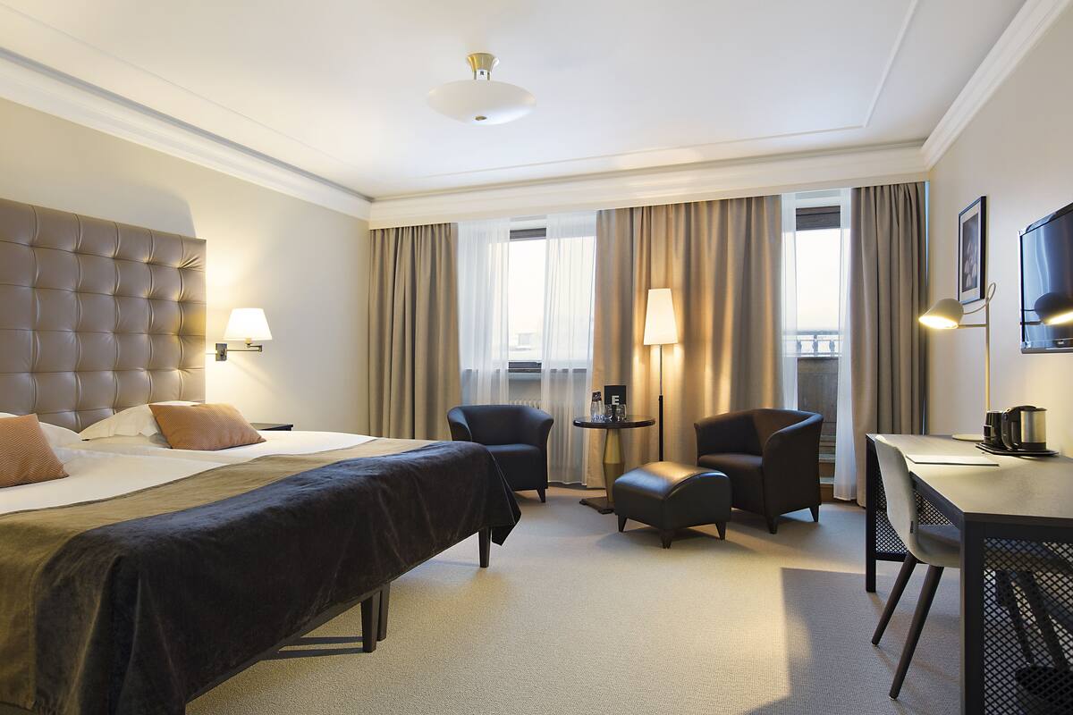 Habitación de hotel con una cama grande, un escritorio, dos sillas, una mesita y una televisión.