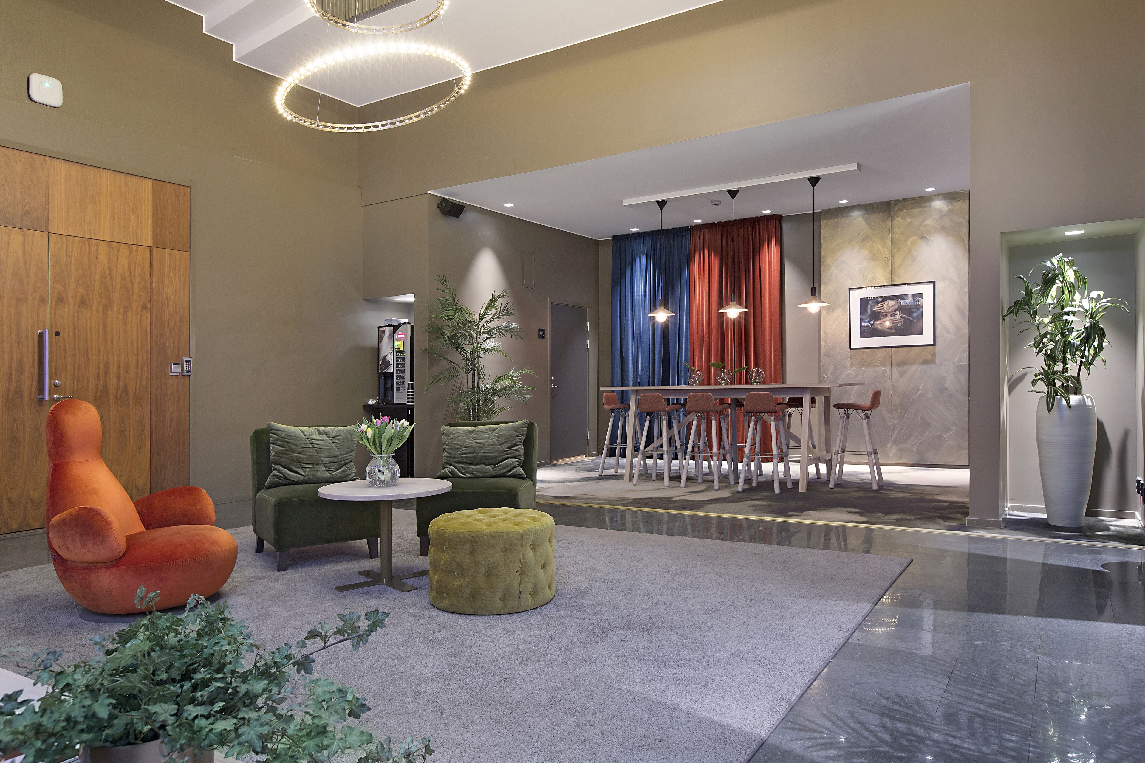 A modern lobby with a wooden wardrobe, a green sofa, an orange chair, a yellow ottoman, a dining area with red curtains, and a large potted plant.