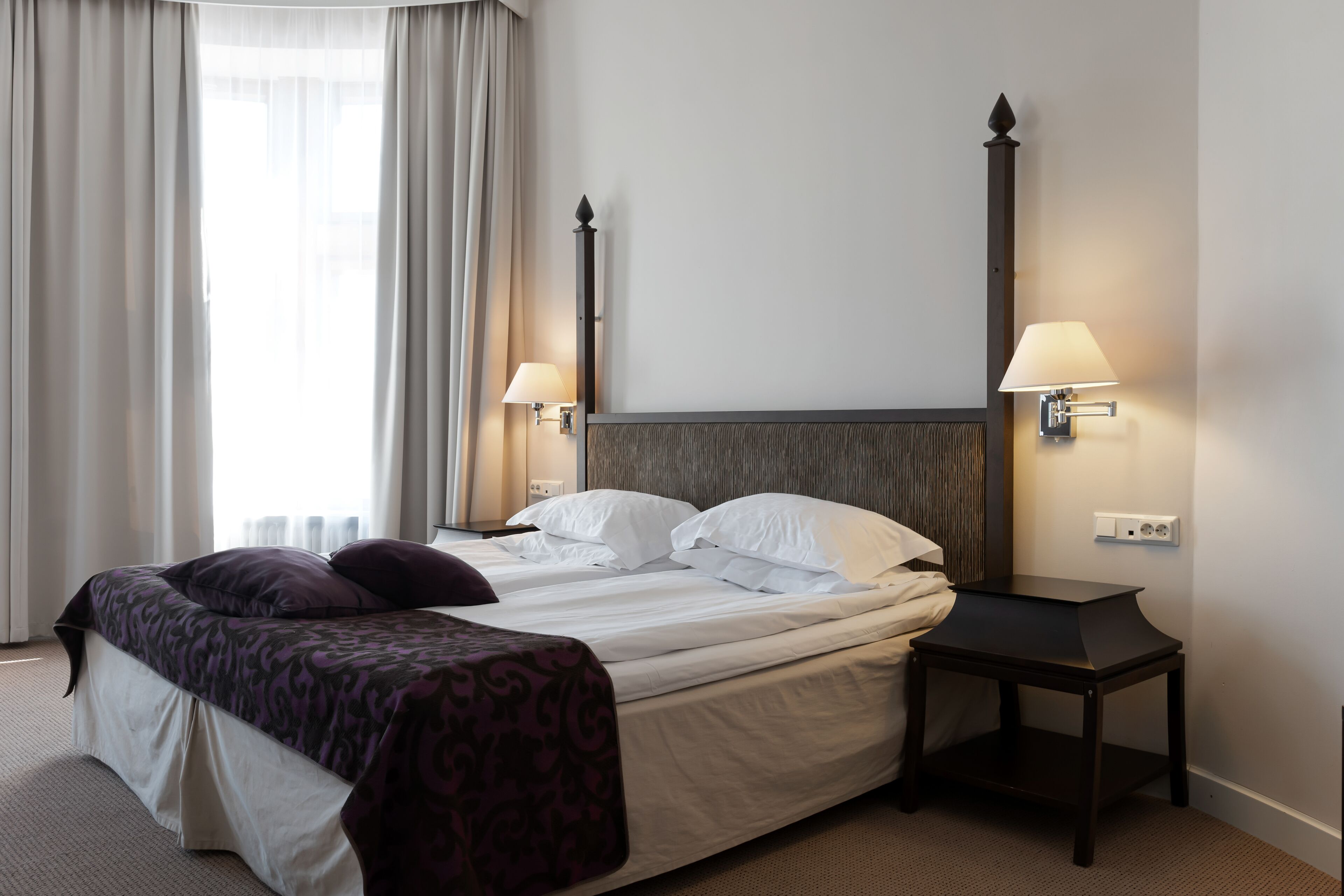 A hotel room with a bed, two bedside lamps, a nightstand, and a window with curtains.