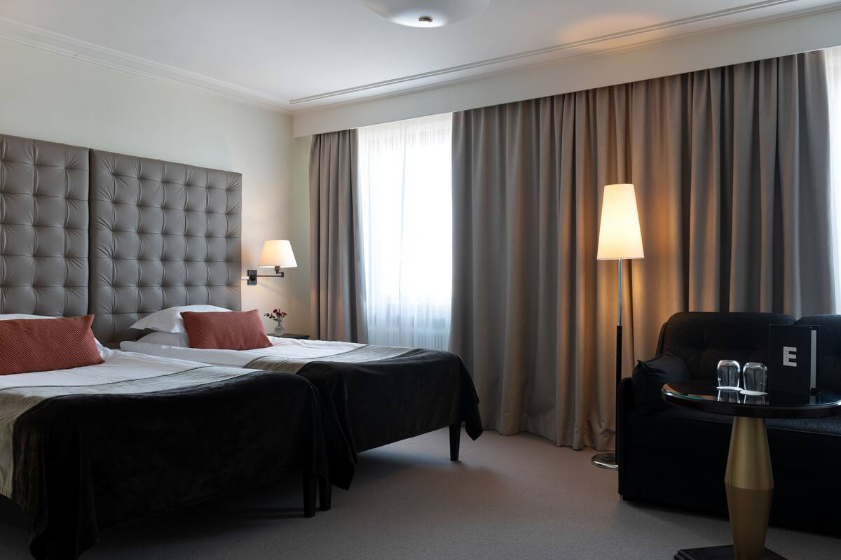 Habitación de hotel con cabecera capitonada, dos camas con ropa de cama roja y blanca, un sofá negro, una mesa redonda con una computadora portátil y una lámpara.