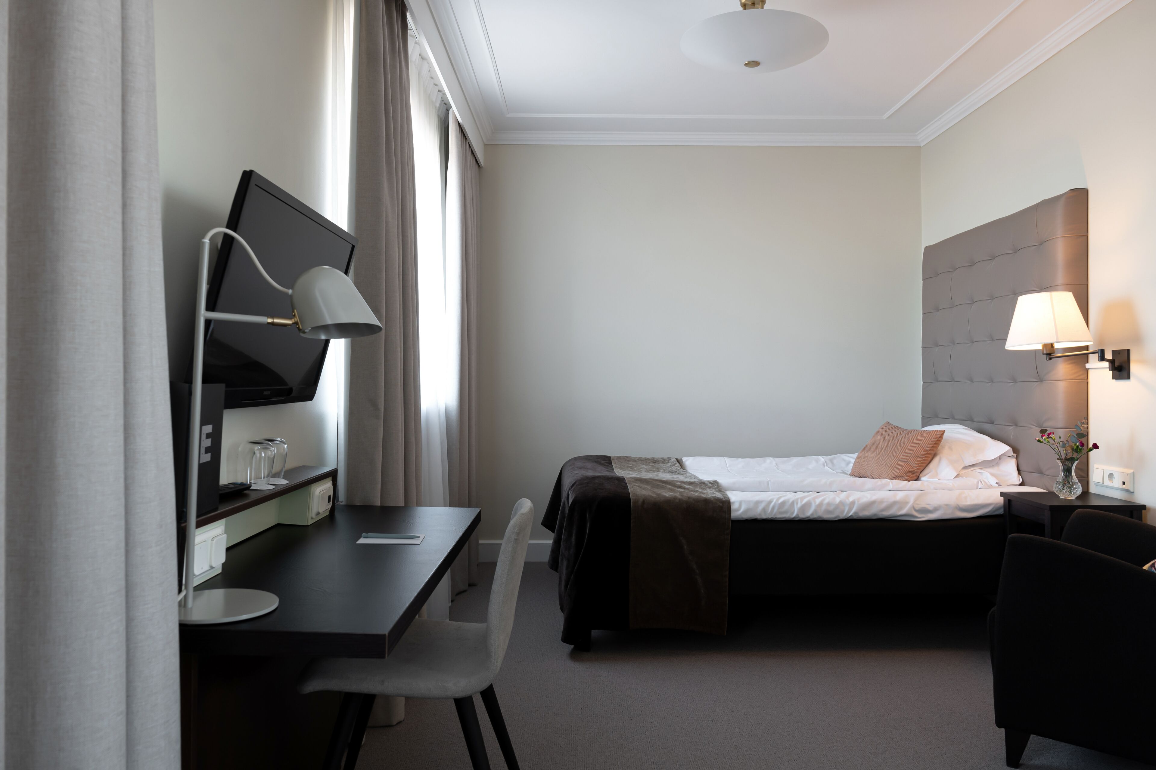 Quarto de hotel com uma cama, uma escrivaninha com computador, uma cadeira, um abajur e uma mesinha de cabeceira com um vaso.