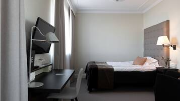 Quarto de hotel com uma cama, uma escrivaninha com computador, uma cadeira, um abajur e uma mesinha de cabeceira com um vaso.