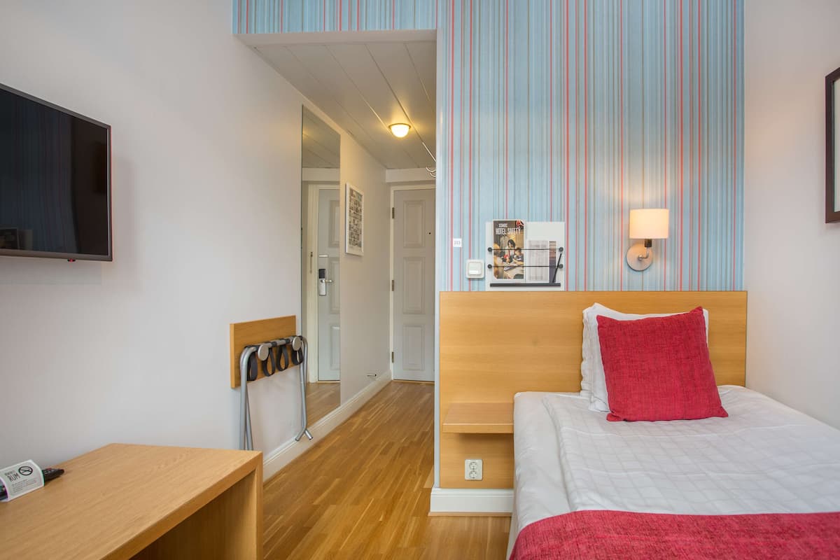 A hotel room with a wooden desk, a bed with a red pillow, a TV mounted on the wall, and a striped wallpaper design.
