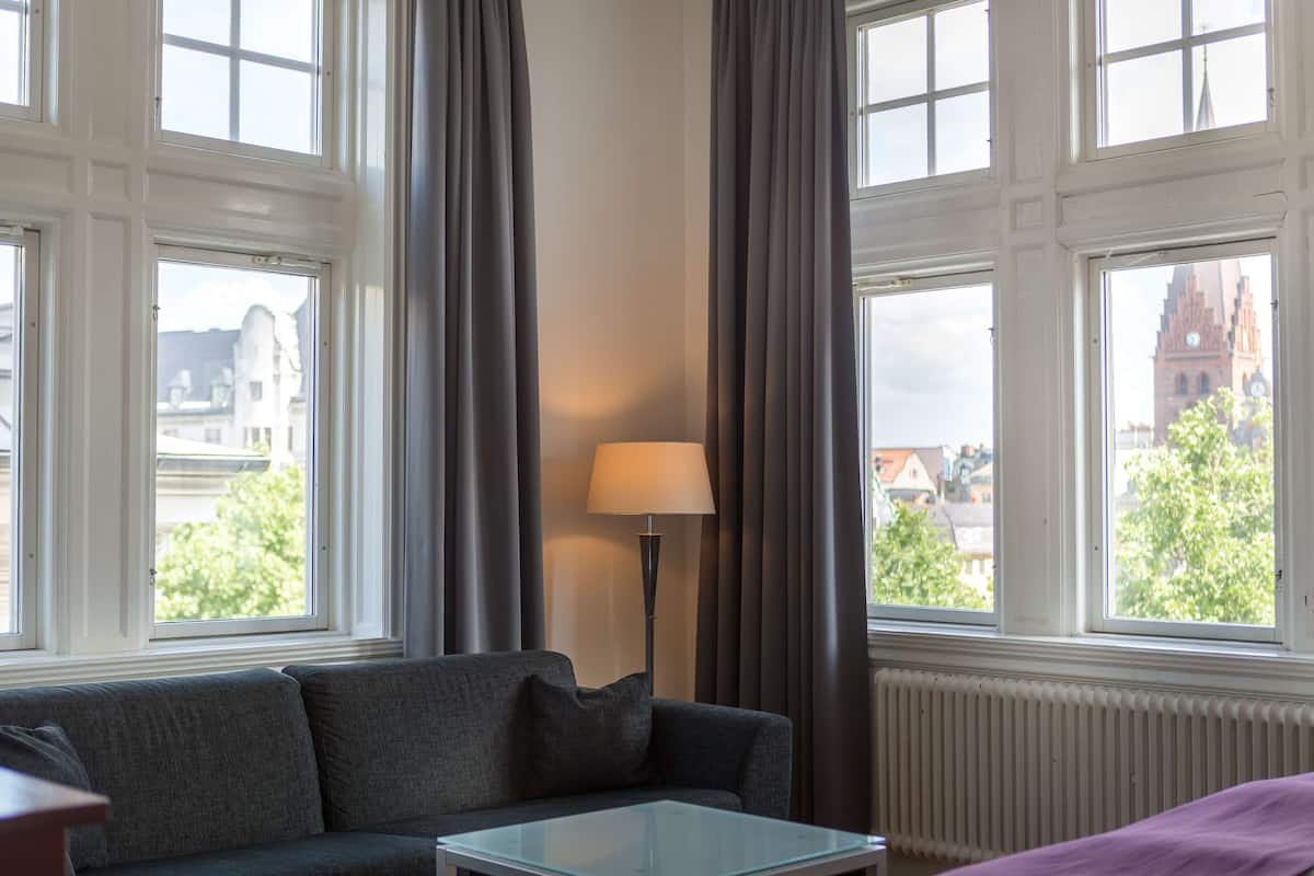 A room with a grey sofa, a glass coffee table, and a lamp. There are large windows with curtains, and a view of buildings and trees outside.