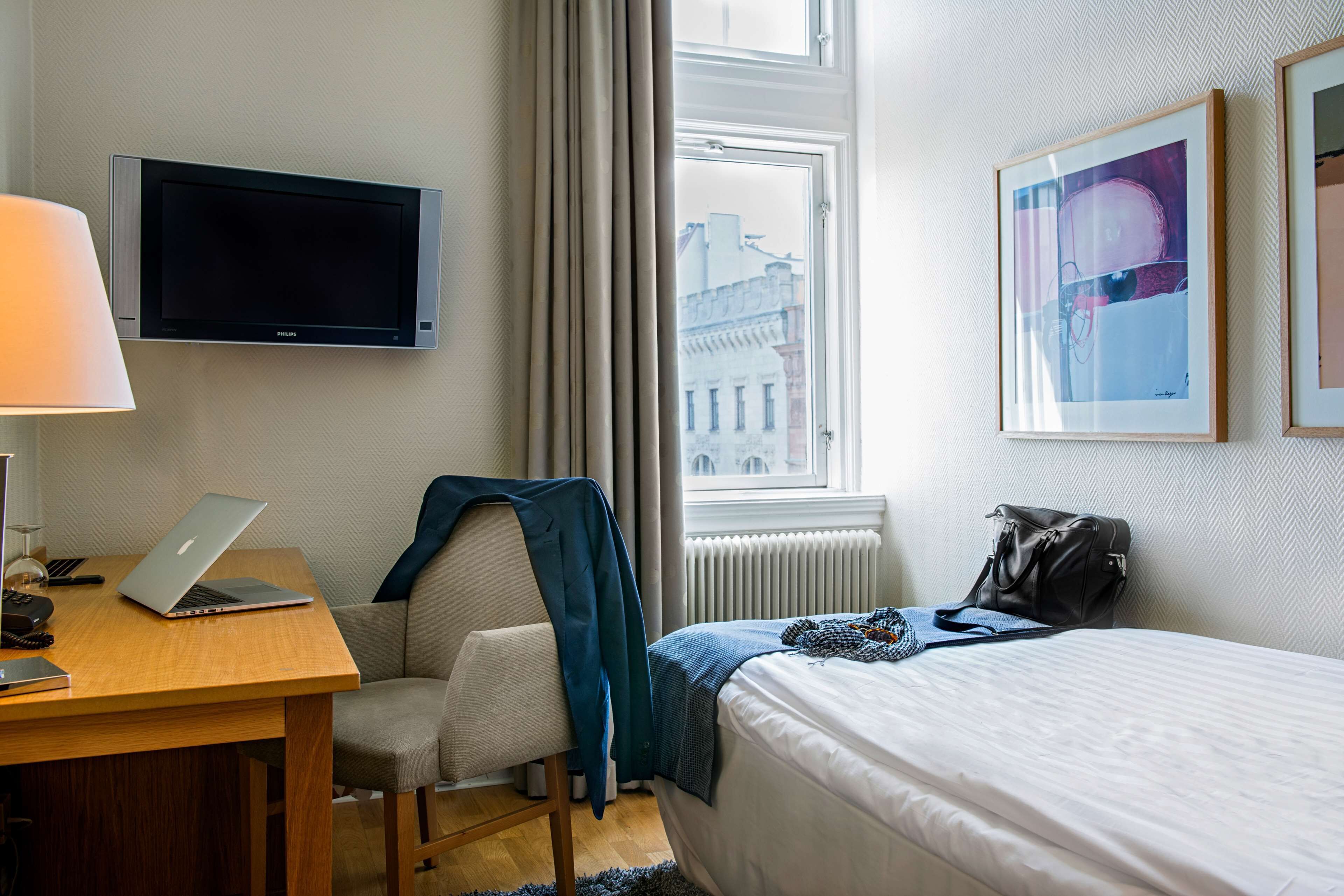A hotel room with a bed, a desk with a laptop, a chair, a TV mounted on the wall, a window with a view of buildings, and a framed picture on the wall.