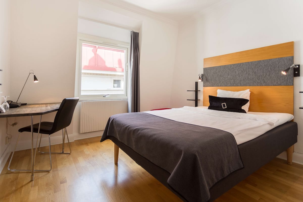 A modern hotel room with a wooden floor, a bed with a grey and white headboard, a desk with a chair, and a window with curtains.