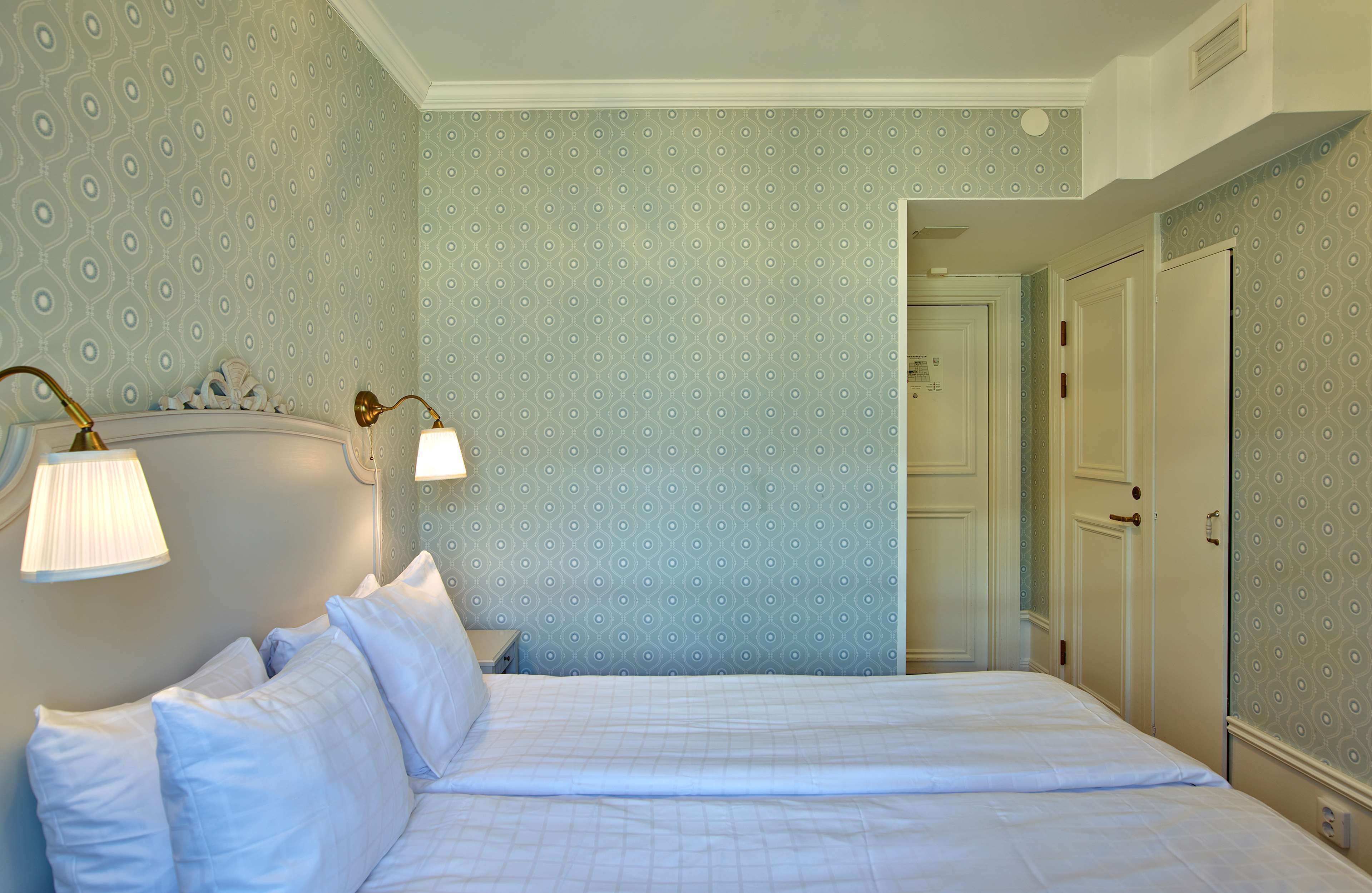 A hotel room with a bed, two bedside lamps, and a patterned wallpaper.