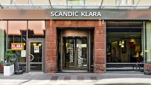 The exterior of Scandic Klara hotel with a glass entrance, a signboard, and a potted plant. - Scandic Klara (Stockholm)