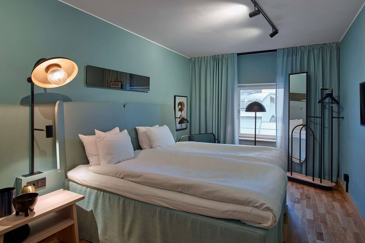 A modern hotel room with a large bed, a bedside lamp, a flat-screen TV, and a view of the sky through the window.