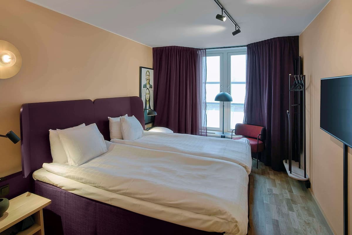 A hotel room with a large bed, a desk, a chair, a TV, and a window with curtains.