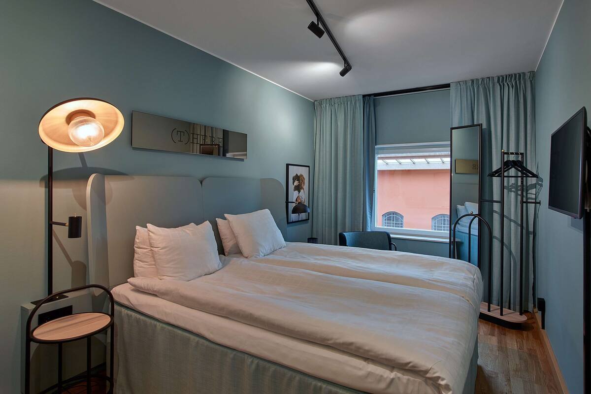 A hotel room with a large bed, a bedside table, a lamp, a TV, and a window with curtains.