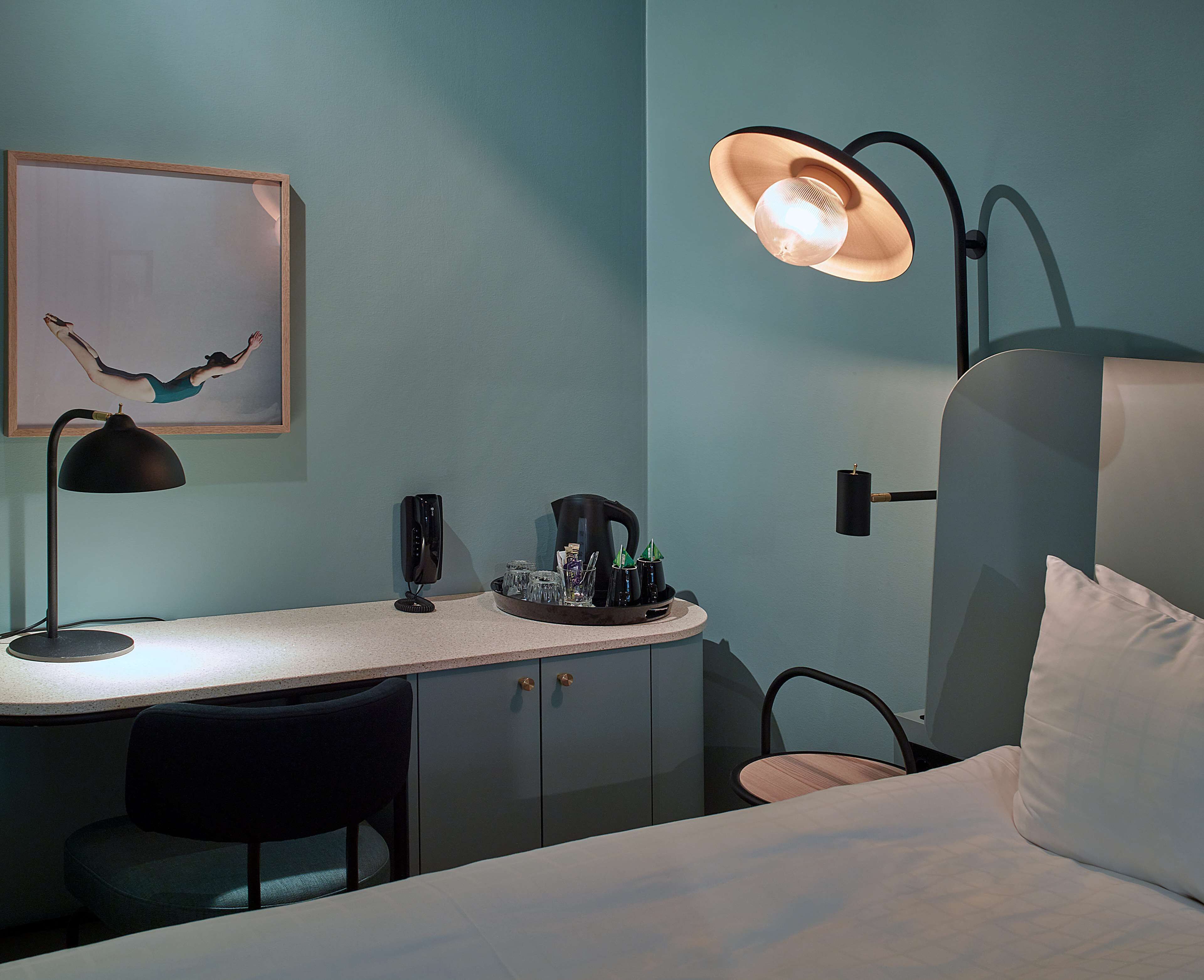 A hotel room with a bed, a desk with a lamp, a chair, a coffee maker, and a framed picture on the wall.
