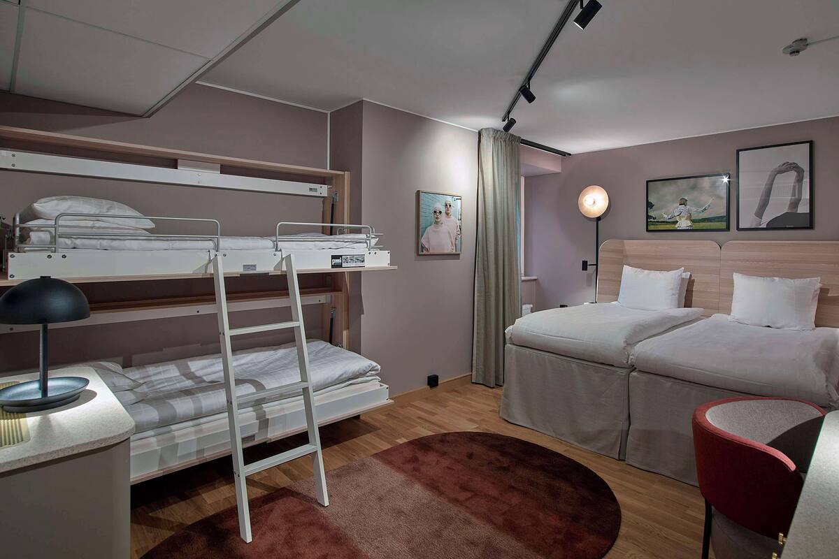 A bedroom with bunk beds, a desk with a lamp, a bedside table, a chair, and a bed with a headboard.