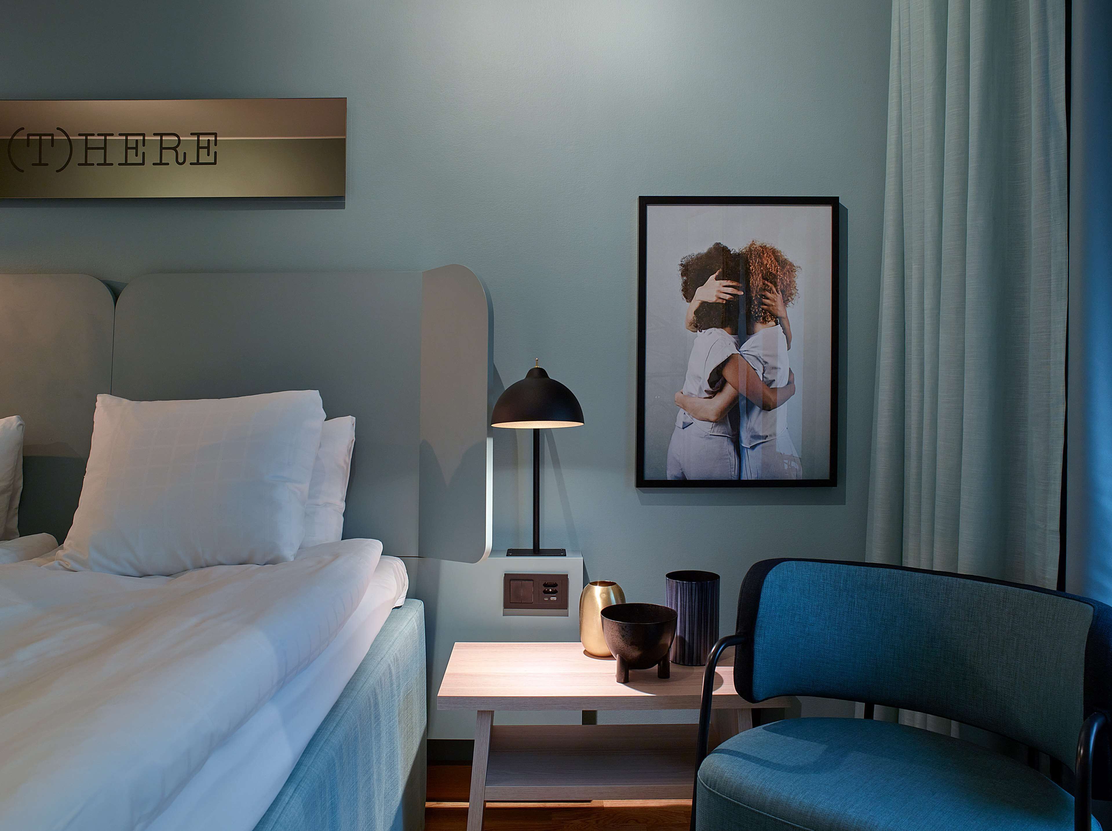 Habitación de hotel con cama, mesita de noche con lámpara, un cuadro enmarcado y una silla.