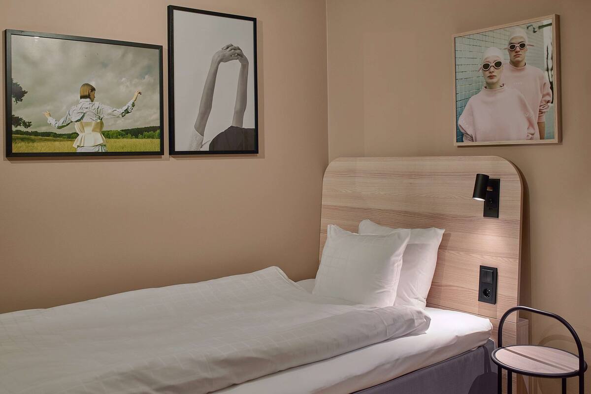 A bedroom with a bed, a nightstand, a wall-mounted lamp, and framed pictures on the wall.