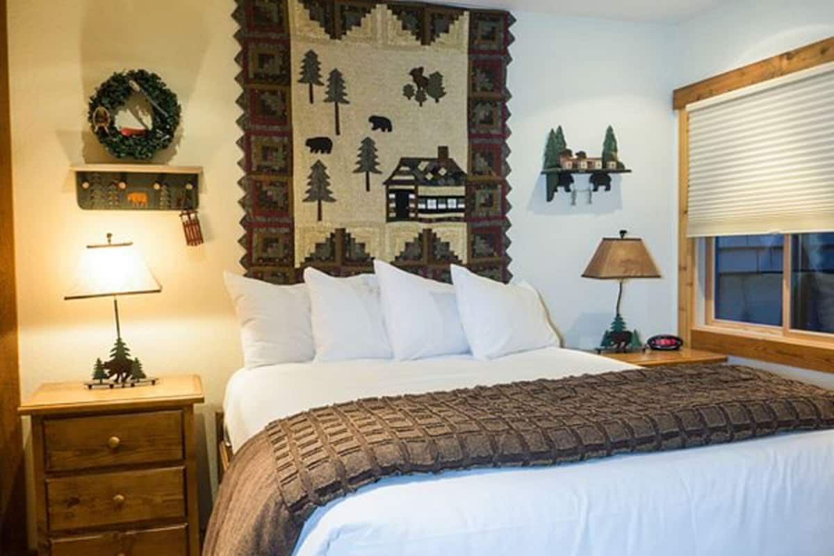 Un dormitorio con cama, mesita de noche con lámpara, cómoda, un adorno en la pared y ventana con persianas.