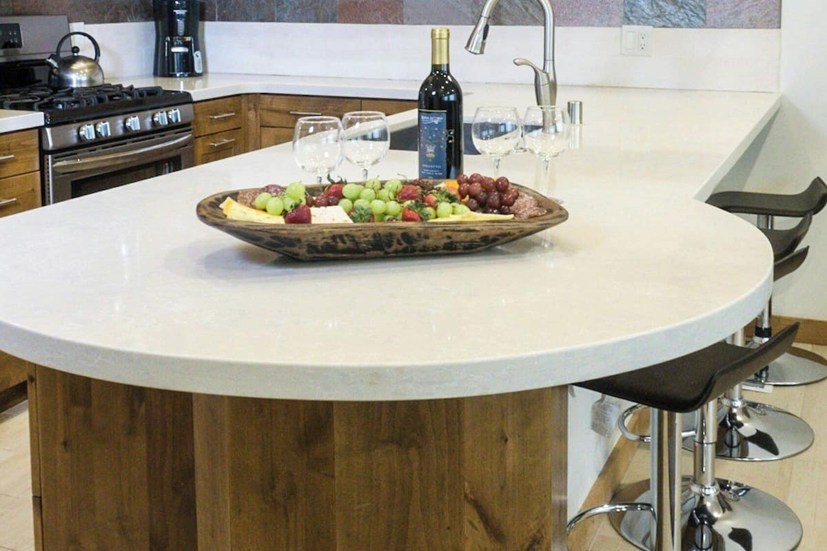 Una cocina con armarios de madera, una isla central con una bandeja de fruta, una botella de vino y taburetes de bar.