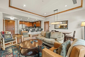 A modern living room with a sofa, coffee table, and chairs. A kitchen is visible in the background with wooden cabinets and a dining area. - Northstar Resort by Vail Resorts (Truckee)
