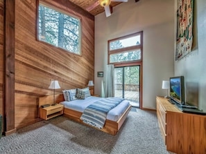 A bedroom with a large bed, two bedside tables, a TV, and a view of the outdoors through a large window. - Northstar Resort by Vail Resorts (Truckee)